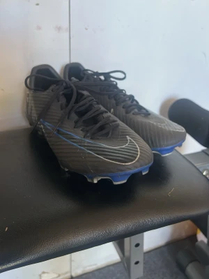 Svarta Nike fotbollsskor med dobbar pro - Säljer ett par svarta fotbollsskor från Nike med vita detaljer och blå sula. Skorna har snörning och slitstarka dobbar för bra grepp på planen. Snygg design med diskreta ränder och klassisk Nike-logga på sidan. Perfekta för dig som vill ha stilrena och funktionella skor.