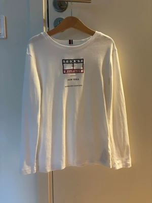 Vit långärmad topp Tommy Hilfiger - Vit långärmad topp från Tommy Hilfiger med tryckt logga och text på bröstet. Klassisk rund halsringning och mjukt material som känns skönt mot huden. Perfekt till jeans eller kjol för en clean och enkel look.