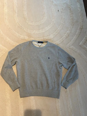 Ralph Lauren tröja grå  - Klassisk grå sweatshirt från Polo Ralph Lauren med rund halsringning och broderad marinblå logga på bröstet. Tröjan har ribbade muddar vid ärmslut och nederkant, perfekt för en avslappnad och stilren look. Skick 9/10