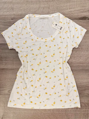 Vit t-shirt med citronmönster XS/S 157 - Fräsch vit t-shirt med små gula citroner över hela plagget. T-shirten har rund halsringning och korta ärmar. Tillverkad i mjuk bomull som känns skön mot huden. Kan användas både som tunn tröja och som pyjamas. Har fortfarande lapp kvar.