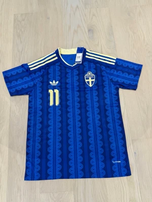 Blå Sverige fotbollströja Adidas med elanga  - Säljer en blå fotbollströja från Adidas med Sveriges landslagsemblem och nummer 11 på bröstet. Tröjan har gula detaljer, klassiska Adidas-ränder på axlarna och ett retroinspirerat mönster. Perfekt för dig som vill stötta Sverige med stil.