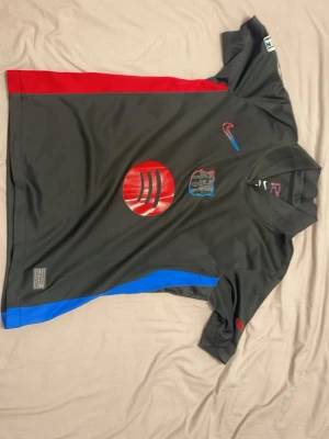 Svart FC Barcelona borta matchtröja Nike Lamine Yamal 24/25 - Säljer en svart Barcelona matchtröja från Nike med röda och blå detaljer. Tröjan har klubbmärke, Nike-logga och sponsortryck på bröstet samt namn och nummer (Lamine Yamal 19) på ryggen. Tillverkad i ett lätt och ventilerande material, perfekt för fotboll.