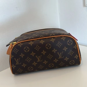 Brun  monogram necessär i klassisk modell och bra storlek  - Säljer en klassisk necessär i brunt med två dragkedjor upptill och guldiga metalldetaljer. Perfekt för att organisera dina essentials på resan eller i vardagen. Nyskick då den aldrig använts. Både praktisk och trendig. 