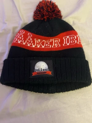 Svart och röd mössa med tofs - Säljer en svart och röd stickad mössa med tofs och uppvikt kant. Mössan har ett broderat märke med texten 'Tvååker Innebandy' och en innerbandy boll på framsidan. Röd rand och text runt om samt matchande tofs på toppen. Perfekt för kalla dagar.