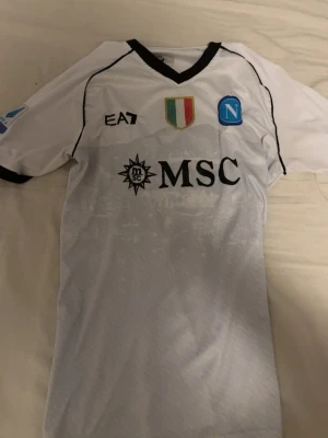 Napoli fotbollströja - Snygg vit matchtröja från Napoli med svarta detaljer och klubbmärke på bröstet. Tröjan har sponsorloggor som MSC och Upbit, samt svarta muddar och V-ringad krage. Perfekt för dig som älskar fotboll och vill bära ditt lag med stil.