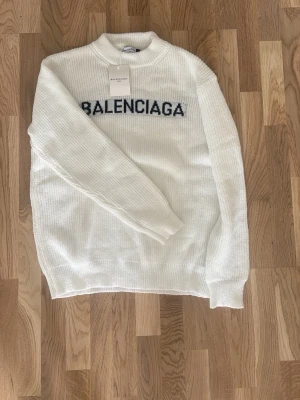 Vit stickad tröja - Vit stickad tröja med svart logotyp över bröstet. Tröjan har rund halsringning och ribbstickade muddar vid ärmslut och nederkant. Perfekt för dig som gillar exklusiva och trendiga plagg. Passar XL