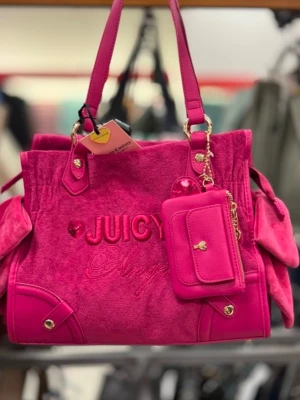 Juicy Couture rosa handväska - Snygg rosa handväska från Juicy Couture i mjukt material med broderad text 'Juicy Angel' på framsidan. Väskan har dekorativa gulddetaljer, sidofickor och matchande liten plånbok med hjärtformad knapp. Perfekt för dig som gillar färgstarka accessoarer.