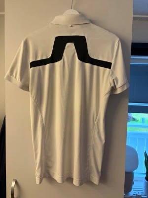 Golfpiké J.Lindeberg  - Säljer en stilren vit pikétröja från J.Lindeberg med svart logga på kragen och en unik svart detalj över ryggen. Klassisk krage och knäppning framtill. Perfekt för dig som gillar sportig och clean stil. Strl S passar M, mycket bra skick men har någon liten defekt som inte syns vid användning 