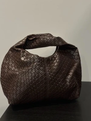 Mörkbrun flätad handväska - Köptes på marknad i Spanien! Snygg mörkbrun handväska i flätad läderimitation med rundad form och mjukt handtag. Väskan har en modern och trendig look som passar perfekt till olika outfits. Perfekt accessoar för dig som gillar stilrena detaljer.