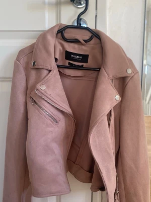 Rosa bikerjacka från Pull&Bear - Snygg rosa skinnjacka i bikerstil från Pull&Bear. Jackan har silvriga dragkedjor, snedställd front och breda slag. Tillverkad i mjukt material med en croppad passform och coola detaljer som axelknappar och ficka med dragkedja.