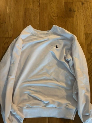 Vit sweatshirt med broderad logga (polo) - Klassisk vit sweatshirt med rund hals och ribbade muddar. Tröjan har en liten broderad logga i svart på bröstet. Perfekt för en clean och stilren look. Passar till både jeans och mjukisbyxor. Pris kan diskuteras vid snabb affär!