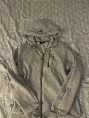 Grå hoodie från Sail Racing - Säljer en grå hoodie från Sail Racing med dragkedja framtill och en bröstficka med dragkedja. Den är ändå i bra skick skulle jag säga, dragkedjan går fortfarande att öppna o stänga även fast den dära grejen inte är kvar. Tröjan var förut rätt sliten nere vid ärmarna men det tog jag bort så nu tänker man inte på det