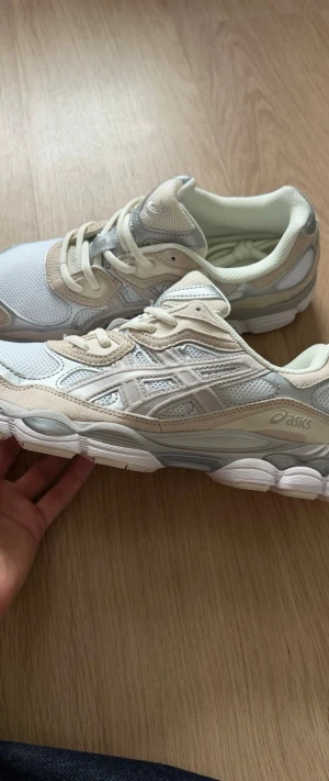 Vita och beige Asics sneakers - Snygga sneakers från Asics i vitt och beige med inslag av grått. Skorna har mesh- och mockadetaljer, klassisk snörning och en chunky sula för extra komfort. Perfekta för dig som gillar stilrena och sportiga vibes.