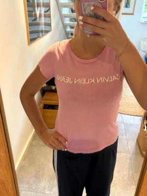 Rosa Calvin Klein T shirt - Snygg rosa t-shirt från Calvin Klein Jeans med vit logga tryckt över bröstet. Modellen är kortärmad och har en klassisk rund halsringning. Perfekt för dig som gillar en enkel men trendig stil.