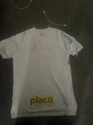 Vit Umbro matchtröja med tryck - Säljer en vit matchtröja santos  från Umbro med tryck på ryggen och ärmen. Tröjan har korta ärmar och reklamtryck i blått och gult, bland annat 'placo' och 'Kicouros'. Perfekt för träning eller match.