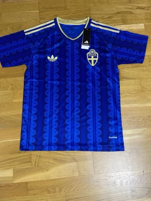 Blå Sverige fotbollströja Adidas - Säljer en blå fotbollströja från Adidas med Sveriges landslagsemblem på bröstet. Tröjan har ett unikt mönster i blå nyanser, vita Adidas-ränder på axlarna och gul krage. Helt ny aldrig använd och kommer med påsen. Unisex funkar för både tjejer och killar 