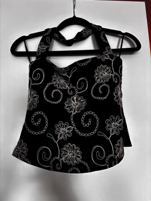 Svart halterneck topp med broderi BIK BOK - Snygg svart halterneck topp med vita broderade blommor och snirkliga mönster. Toppen har dragkedja i sidan och klister vid byst så att den hålls uppe och ej glider ner. Har inte kommit till användning tyvärr trots skitsnygg!