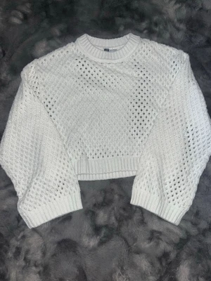 Vit cropped stickad tröja - Säljer en vit cropped stickad tröja med hålmönster och rund hals. Tröjan har långa ärmar och ribbade muddar vid ärmslut och nederkant. Perfekt för lager på lager och ger en trendig look till din outfit.
