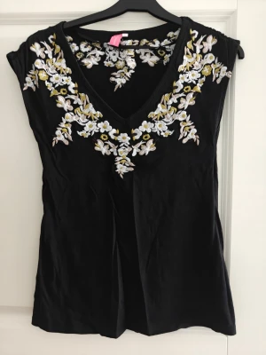 Svart blommig topp från 365 Sunshine - Svart topp från 365 Sunshine med v-ringning och broderade blommor i vitt och gult runt halsen. Ärmlös modell med mjukt material och figurnära passform. Perfekt för dig som vill ha en söt och unik detalj i garderoben.köpt på MQ