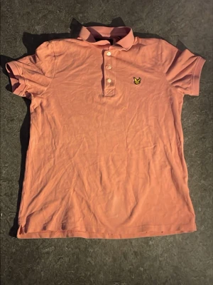 Rosa pikétröja med krage - Snygg rosa pikétröja med klassisk krage och knappar framtill. Tröjan har korta ärmar och ett broderat gult emblem på bröstet. Tillverkad i bomull för en bekväm känsla. Perfekt till jeans eller shorts för en avslappnad look.