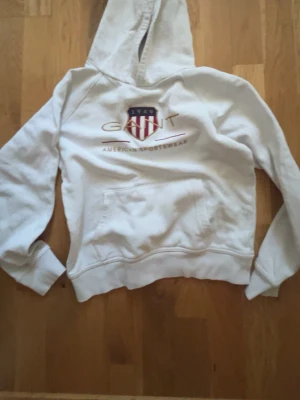 Vit hoodie från GANT med tryck - Vit hoodie från GANT med broderad logga och sköld på bröstet samt texten 'American Sportswear'. Klassisk känguruficka framtill och ribbade muddar. Perfekt för en avslappnad och sportig stil.