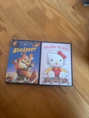 DVD-filmer: Kogänget & Hello Kitty - Två DVD-filmer: Disneys Kogänget med färgglad tecknad framsida och Hello Kitty Stubbestad 1 med söt Hello Kitty-figur på omslaget. Båda fodralen är svarta och gjorda av plast, perfekta för dig som gillar animerade filmer och samlar på barnfilmer.