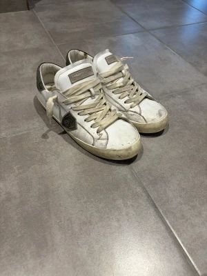 Vita Philippe Model sneakers - Säljer ett par vita Philippe Model sneakers med beigea skosnören och grå detaljer. Skorna har en klassisk låg profil och en logotyp på sidan. Tillverkade i läder med en robust gummisula. Perfekta för dig som gillar streetwear och stilrena detaljer.