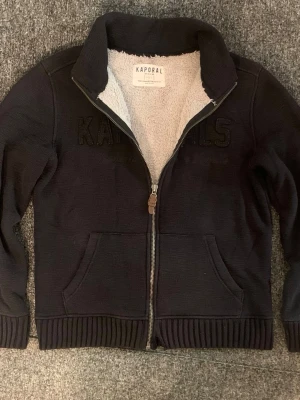 Svart ziptröja från Kaporal - Mysig svart ziptröja från Kaporal med broderad logga på bröstet. Tröjan har mjukt vitt teddyfoder på insidan, två fickor framtill och ribbade muddar vid ärmslut och nederkant. Perfekt för kyliga dagar och riktigt skön att ha på sig.