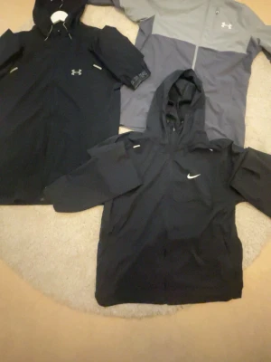 Svarta och grå vindjackor Nike & Under Armour - Tre sportiga vindjackor, två svarta och en grå/svart, från Nike och Under Armour. Alla har huva och dragkedja framtill. Perfekta för blåsiga dagar och har en clean, modern look med logga på bröstet.