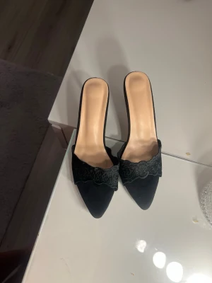 Svarta spetsiga pumps med spetsdetalj - Säljer ett par svarta pumps med öppen häl och spetsig tå. Skorna har en elegant spetsdetalj över foten och smal klack. Materialet ser ut att vara mockaimitation med inslag av spets, vilket ger en lyxig känsla. Perfekta för dig som vill ha en chic och trendig look. Storlek 39. 