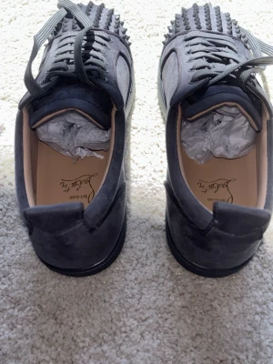 Gråa Louboutin sneakers med nitar - Säljer ett par gråa sneakers från Christian Louboutin med ikoniska röda sulor och coola nitar på tån. Skorna är låga och har snörning samt en lyxig känsla med mockaliknande material. Perfekta för dig som vill sticka ut med din stil. NYTT SKICK ALDRIG ANVÄNT passar 44 pris kan diskuteras hör av er om ni har frågor! PRIS KAN DISKUTERAS 