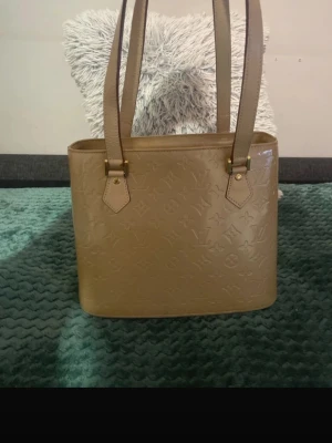 Beige handväska i lackat läder - Snygg beige handväska i lackat läder med präglat monogrammönster och två långa axelband. Väskan har guldfärgade metalldetaljer och dragkedja upptill. Perfekt för dig som gillar stilrena accessoarer med lyxig känsla.