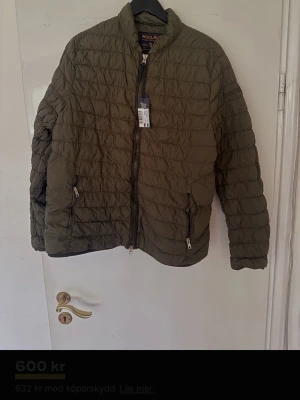 Olivgrön pufferjacka från Woolrich - Snygg olivgrön pufferjacka från Woolrich med quiltad design och dragkedja framtill. Jackan har två fickor med dragkedja och står upp lite i kragen. Perfekt för kalla vinterdagar och riktigt skön att bära.
