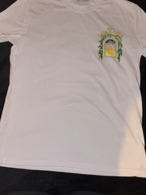 Vit t-shirt med tryck Casablanca - Säljer en vit t-shirt med färgglatt tryck av ett fönster och texten 'Avenida Casablanca' både fram och stort på ryggen. T-shirten har rund hals och korta ärmar. Perfekt för dig som gillar grafiska motiv och stilrena plagg.