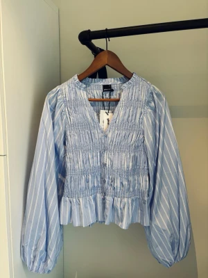 Blå- och vitrandig blus med v-ringning - Prislappen kvar! Snygg blå- och vitrandig blus med v-ringning och smockdetaljer över fram och baksida. Blusen har långa puffärmar och en lätt volang nedtill. Tillverkad i ett ganska tunt material som känns luftigt och fräscht. Nypris 399kr