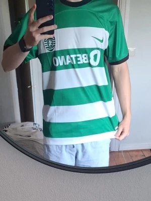 Grönvit randig t-shirt SUPER BOCK - Säljer en grön och vit randig t-shirt med texten SUPER BOCK på ryggen. T-shirten har klassisk passform och korta ärmar. Perfekt för dig som gillar sportig och avslappnad stil.