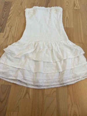 Vit axelbandslös volangklänning - Söt vit axelbandslös klänning med smock upptill och dubbla volanglager nedtill. Klänningen har broderade små blommor i tyget och är perfekt för sommarens varna dagar. Klänningen har en fläck i höger och vänster topp på klänningen och den har även två sydda hål som man lätt kan sy ihop igen. De hålen fans när den köptes. ENDAST ANVÄNT EN GÅNG!!