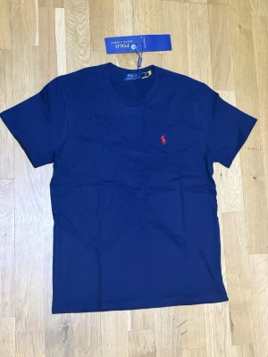 Marinblå t-shirt från Polo Ralph Lauren - Snygg marinblå t-shirt från Polo Ralph Lauren med klassisk röd logga broderad på bröstet. T-shirten har rund halsringning och korta ärmar. Tillverkad i mjuk bomull för en skön känsla.