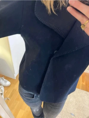 Mörkblå kappa med bred krage - Stilren mörkblå kappa med bred, markerad krage och rak passform. Kappan har en minimalistisk design utan synliga knappar och är tillverkad i ett tjockare tyg som passar perfekt för kyligare dagar. Inte min bild men kan skicka fler bilder vid intresse.