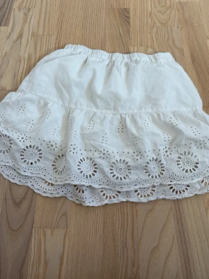 Vit kortkjol med broderade spetsdetaljer - Söt vit kortkjol med bred volangkant och broderade spetsdetaljer i blommigt mönster. Kjolen har resår i midjan och är gjord i ett lätt bomullstyg, perfekt för varma dagar. Den har två super små fläckar på framsidan med dom syns knappt vid användning!