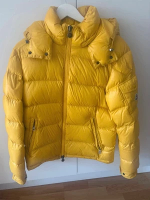 Gul moncler Maya jacka  - Snygg gul dunjacka från Moncler med glansig finish, hög krage och avtagbar huva. Jackan har dragkedja framtill, två fickor med dragkedja och en ficka med lock på ärmen med Moncler-logga. Perfekt för kalla dagar och riktigt trendig stil. Några svarta fläckar som syns på bilden men borde gå att tvätta bort men har inte försökt. Jacka är i storlek 2 som motsvarar typ medium