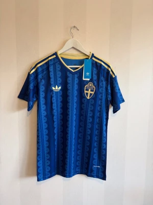 Blå Sverige fotbollströja Adidas - Säljer en blå fotbollströja från Adidas med Sveriges landslagsemblem på bröstet. Tröjan har gula detaljer, mönstrade ränder och klassiska Adidas-ränder på axlarna. Perfekt för match eller träning. Materialet är lätt och andas bra.