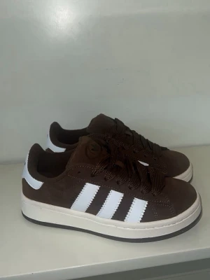 Bruna Addidas campus - Snygga bruna sneakers från Adidas Campus  med klassiska vita ränder på sidorna och vit sula. Skorna har lågt skaft, bruna skosnören och är tillverkade i mockaliknande material. Perfekta för dig som vill ha en clean och sportig look. storlek 36