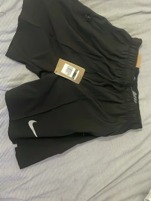 Svarta träningsshorts från Nike - Snygga svarta träningsshorts från Nike med elastisk midja och diskret Nike-logga på benet. Tillverkade i lätt och funktionellt material som passar perfekt för löpning eller gym. Klassisk design med normal passform och enkel look.