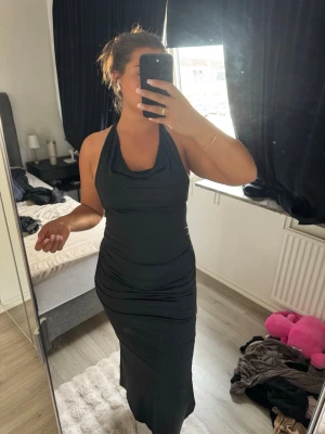 Svart långklänning med halterneck - Stilren svart långklänning med halterneck och draperad urringning. Klänningen har en figurnära passform och är ärmlös, vilket ger en elegant och modern look. Perfekt för dig som vill ha en klassisk och tidlös stil.