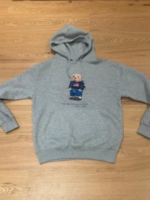 Polo bear hoodie  - Säljer en grå hoodie med huva och magficka. På bröstet finns ett tryck av en björn i blå outfit och amerikansk flagga. Hoodien har ribbade muddar vid ärmar och nederkant. Perfekt för dig som gillar streetwear och vill ha något bekvämt och stilrent.