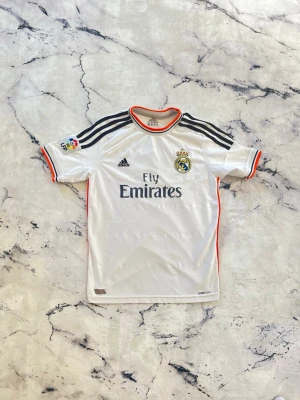 Real Madrid Ronaldo matchtröja - Vit Real Madrid matchtröja från Adidas med svarta ränder på axlarna, orange detaljer och klubbmärke på bröstet. Tryck med 'Fly Emirates' fram och 'Ronaldo 7' bak. Korta ärmar och sportig passform, perfekt för fotbollsträning eller support.