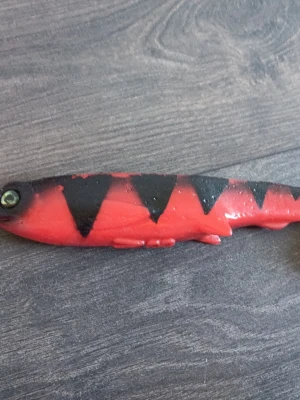 Röd och svart fiskjigg - En fiskjigg i mjukt plastmaterial, formad som en fisk med röd kropp och svarta triangulära mönster längs ryggen. Jiggen har realistiskt öga och detaljerade fenor, perfekt för fiske efter rovfisk. Hemmagjord 