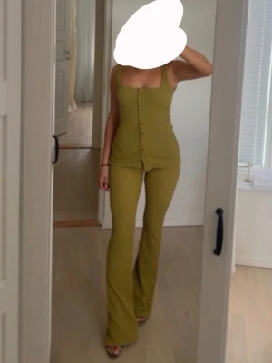 Grönt ribbat set med knappar - Säljer ett grönt ribbat set med linne och matchande byxor. Linnet har breda axelband, fyrkantig urringning och knappar framtill. Byxorna är höga i midjan och har utsvängda ben. Materialet ser stretchigt och bekvämt ut, perfekt för en trendig look.