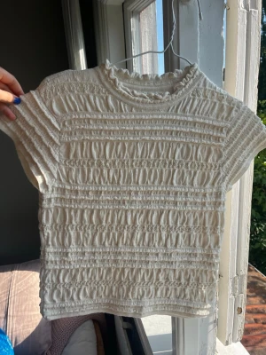 Vit ribbad topp med volangkrage - Supersöt vit topp med ribbad struktur och volangdetalj vid kragen. Kortärmad modell med stretchigt material och fina rynkade partier över hela toppen. Perfekt att styla till jeans eller kjol för en trendig look.